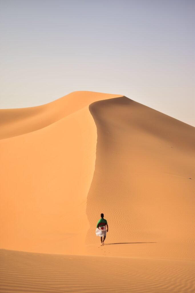 A lone traveler walking over the vast, golden sand dunes of the Sahara Desert in Algeria.